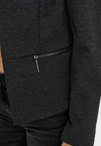 Černá texturovaná bunda s boční kapsou na zip, která má elegantní design a upravený střih. Viditelné detaily šití.