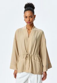 Beige Jacke im lockeren Schnitt mit Wickeldesign, weiten Ärmeln, elastischem Bund und Vordertaschen. Glatte Textur und minimale Nähdetails.