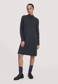 Eine Frau trägt ein schwarzes, knielanges Langarmkleid mit hohem Kragen, kombiniert mit dunkelblauen Socken und schwarzen Slippers, und steht vor einem weißen Hintergrund.