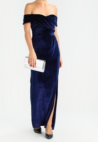 Marinblå off-shoulder maxiklänning i sammet med en slits, som har en figurnära bodice och en flödande kjol. Kombinerat med en silverclutch och gröna klackar.