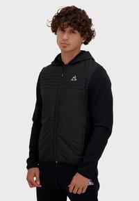 Chaqueta negra con cremallera y un frente acolchado, mangas lisas y capucha. Presenta costuras horizontales y un logo en el pecho. Textura suave.