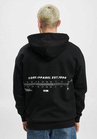 Svart hettegenser laget av en bomullsblanding, med et stort hvitt grafisk trykk av et linjal og teksten "CORE APPAREL EST.1990" på baksiden.
