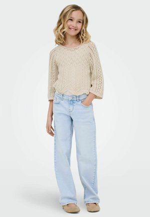 Fată zâmbitoare cu păr blond creț purtând un pulover bej tricotat, blugi bleu deschis și pantofi bej, stând cu o mână în buzunar.