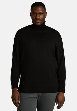 Homme à la peau foncée portant un pull à col roulé noir et un pantalon gris, debout devant un fond blanc uni.