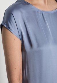 Top di raso azzurro chiaro con una texture liscia, maniche corte e scollatura rotonda. Il tessuto si drappeggia delicatamente sulla spalla e sul braccio.