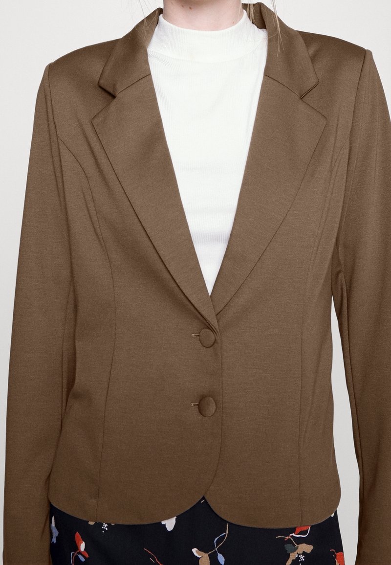 Femme portant un blazer marron à deux boutons sur un top blanc côtelé à col haut et un pantalon sombre à fleurs, visible des épaules à la taille.