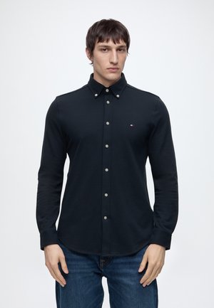 Jeune homme faisant face, portant une chemise noire à boutons avec un petit logo sur la poitrine et un jean bleu sur un fond clair uni.