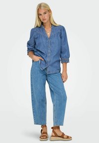 Camicia in denim con bottoni, maniche a sbuffo e polsini arricciati, abbinata a jeans in denim blu a vestibilità ampia e sandali marroni con cinturini.