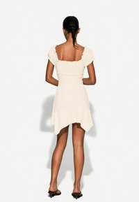 Kleid in heller Cremefarbe mit Puffärmeln, smockiertem Oberteil und asymmetrischem Saum. Textilstruktur mit weichem Finish. Getragen mit schwarzen Sandalen.