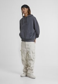 Felpa oversize grigia scura con tasca frontale, abbinata a pantaloni cargo beige con tasche laterali e sneakers chiare.