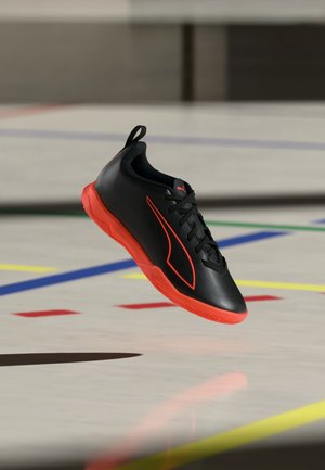 Musta sisäkengän urheilukenkä punaisella pohjalla ja yksityiskohdilla, nauhat solmittu, leijuu kentän lattian yläpuolella, joka on merkitty värikkäillä viivoilla.
