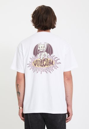 Homme portant un t-shirt blanc avec un graphisme de chat ailé de dragon et le texte "VOLCOM" en police stylisée au dos, sur un fond uni.