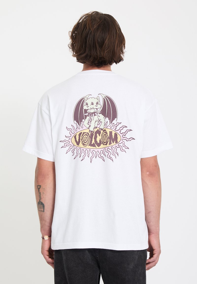 Homme portant un t-shirt blanc avec un graphisme de chat ailé de dragon et le texte "VOLCOM" en police stylisée au dos, sur un fond uni.