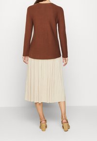 Pull marron à manches longues côtelé associé à une jupe plissée crème. La tenue comprend des sandales à talons beiges et une coupe décontractée.