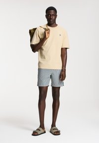 Beige puuvillainen t-paita, raidalliset shortsit ja oliivinvihreä reppä. Sandalit ovat kaksivöiset ja matalapohjaiset. Minimalistinen tyyli.