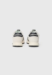 Baskets beiges et noires avec un dessus texturé, dotées d'un col rembourré et d'un logo "CONVERSE" au niveau du talon. Semelle en caoutchouc avec motif de traction.