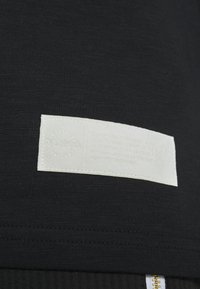 Vit rektangulär tygetikett med upphöjd punktskrift och "PUMA" på svart stickad tyg nära dragkedjan.