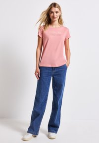 Vaaleanpunainen puuvillateeppari, jossa on käännetyt hihat ja "ENDLESS DREAM" -teksti, yhdistettynä sinisiin leveälahkeisiin farkkuihin ja valkoisiin lenkkareihin.