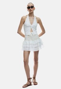 Vestido blanco sin mangas con un cuerpo anudado, que presenta una parte superior texturizada y una falda en capas con volantes; combinado con sandalias marrones de tiras.