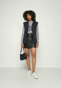 Colete acolchoado azul-marinho sobre uma blusa de mangas longas floral, combinado com uma saia mini de couro preta e ténis brancos. Os acessórios incluem uma mala preta.