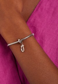 Bracelet en argent ornée d'un design élégant et enroulé, avec un petit pendentif « J » et un accent en diamant, porté sur un bras brun devant un fond rose.