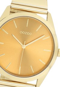 Montre en métal doré avec un cadran doré brossé. Présente des index horaires et des aiguilles minimalistes. Assure lisibilité et élégance dans le design.