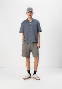 Chemise à manches courtes, bleu clair, avec une poche poitrine, associée à un short gris. Complétée par une casquette grise à motifs et des baskets blanches.