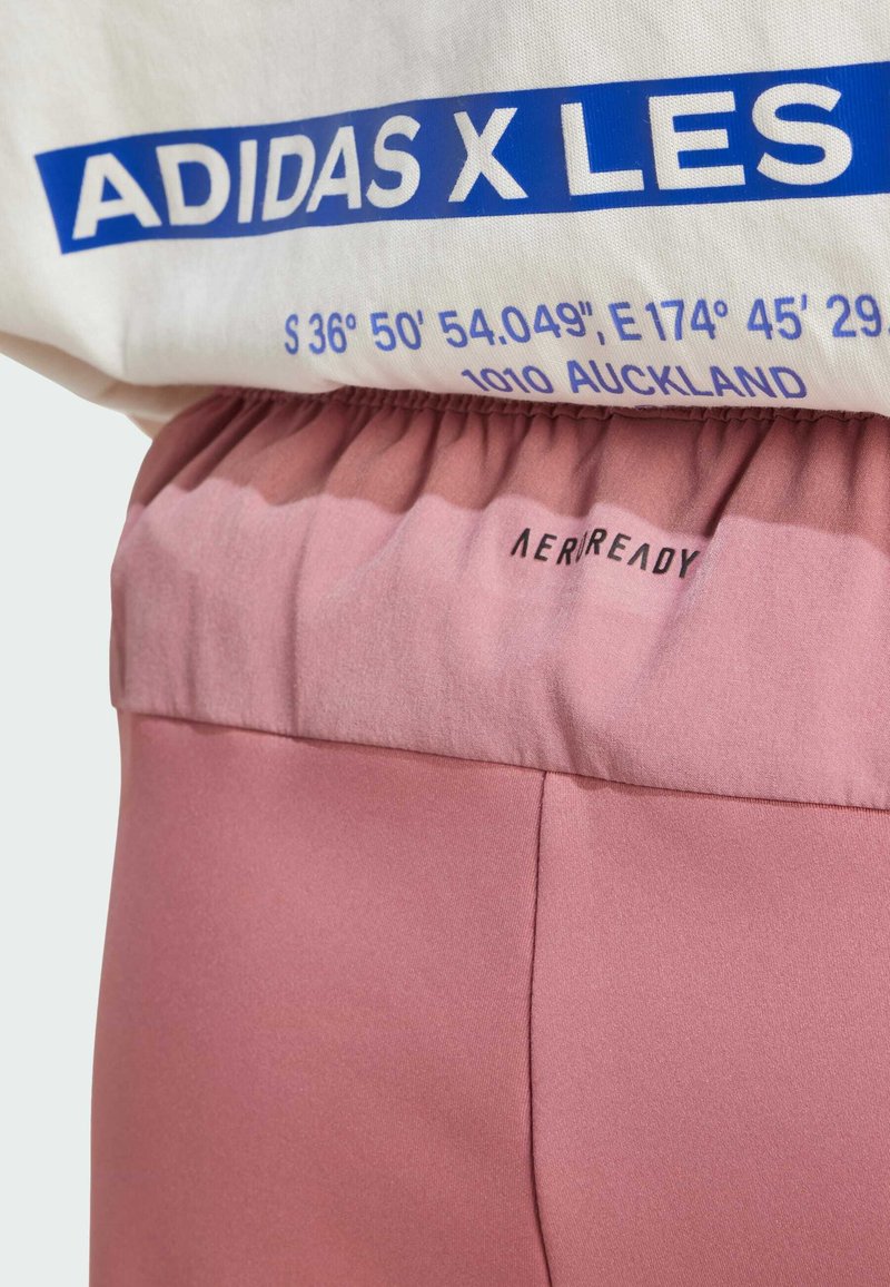 Weißes Oberteil mit blauer Schrift über einem pinken elastischen Bund, der mit "AEROREADY" beschriftet ist. Das Material scheint glatt mit subtiler Textur zu sein.