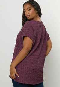 Fioletowa bluzka o fakturowanej strukturze, z krótkimi rękawami i luźnym krojem, z wypukłym, tkanym wzorem oraz zaokrąglonym tyłem.