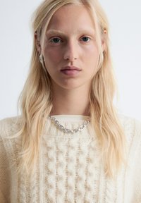 Cremiger Kabelstrickpullover mit einem strukturierten Muster, kombiniert mit einer dicken silbernen Halskette. Lange hellblonde Haare rahmen das Gesicht.