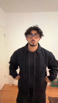 Casaco leve preto com fecho éclair, usado sobre uma camisa preta; óculos grandes e transparentes; cabelo ondulado de comprimento médio; fundo simples.