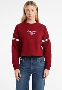 Piros, cropped pulóver fehér hímzett "Guess Jeans" logóval, bordás nyakkivágással és csíkozott részletekkel az ujjain. Kék farmernadrággal párosítva.