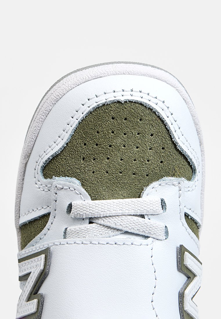 Witte sneaker met olijfgroene suède accenten. Heeft een geperforeerde neusdoos, een gestructureerde bovenkant en grijze veters. Opvallend "N" logo aan de zijkant.