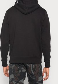 Felpa nera con una leggera texture, dotata di cappuccio regolabile e polsini a costine. Vista da dietro, indossata con pantaloni a camuffamento a fantasia.