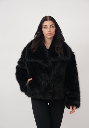 Jonge vrouw met lang donker haar, gekleed in een zwarte nepbontjas en zwarte broek, tegen een effen lichte achtergrond.