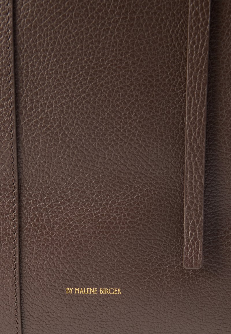 Sac fourre-tout en cuir marron avec une surface texturée, des anses fines et un texte doré embossé. Design simple et fonctionnel avec une apparence classique.