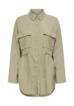 Chemise boutonnee beige clair à manches longues, avec deux poches avant, un détail de taille cintrée et un ourlet arrondi. Texture de tissu lisse.