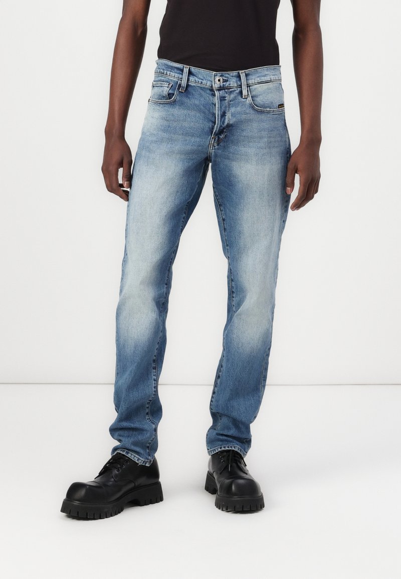 Light blue denim jeans with a straight fit, five-pocket design, contrasting stitching, and wear marks, paired with black chunky shoes.