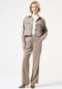 Korte beige ribfluwelen jas met knopenfront en twee borstzakken, gecombineerd met bijpassende ribfluwelen broek en gestructureerde bruine hakken.