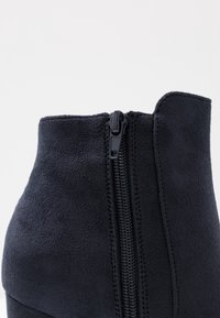 Close-up of a black suede ankle boot with a centered black zipper and visible stitching on a white background.