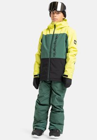 Junge trägt eine gelb-grün-schwarze Schneejacke, grüne Schneehose, schwarze Handschuhe, schwarze Stiefel, eine schwarze Strickmütze und Skibrille und steht.