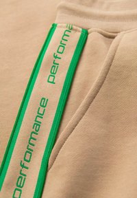 Beige mikina s zeleným pruhem a textem "performance". Hladká textura, ležérní design a šité detaily u kapsy.