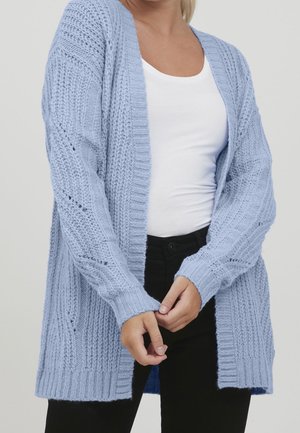 Femme portant un cardigan épais tricoté bleu clair sur une chemise blanche et un pantalon noir, les mains jointes devant.