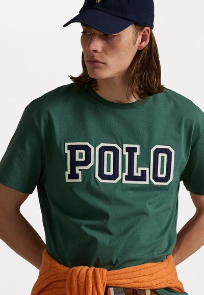 Groene katoenen t-shirt met "POLO" in grote geborduurde letters. Accentuatie met een oranje gebreide trui om de taille geknoopt.
