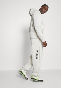 Hvid sweatshirts og matchende bukser med "PARIS" grafik, der har sidelommer og kontrasterende grønne accenter på sneakers.