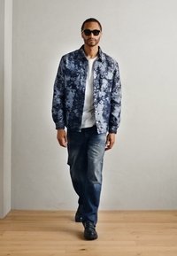 Blue camouflage jacket over white shirt, paired with blue jeans and black shoes. Model wearing black sunglasses, standing on wooden floor.