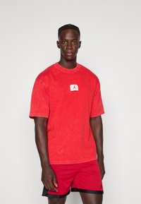 Mand iført en rød vaske-tee med et lille logo-patch og røde basketballshorts med sort kant, stående foran en ensfarvet hvid baggrund.