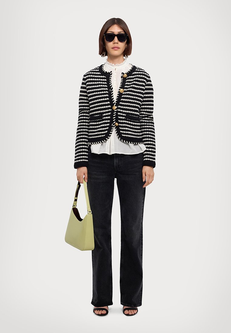Cardigan rayé noir et blanc avec des boutons dorés, superposé sur un chemisier à volants blanc, associé à un jean foncé évasé et un sac à main vert clair.
