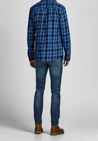 Niebieska koszula flanelowa w kratę z długimi rękawami i plisą z tyłu, zestawiona z dopasowanymi niebieskimi dżinsami i brązowymi skórzanymi butami.