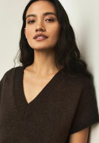 Brun strikket trøje med V-hals og korte ærmer. Blød tekstur og afslappet pasform. Ribstrikkede detaljer langs halsudskæringen.